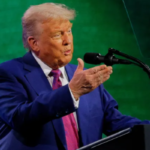 Donald Trump in a blue suit speaking into a microphone, gesturing with his right hand on stage with a green background.