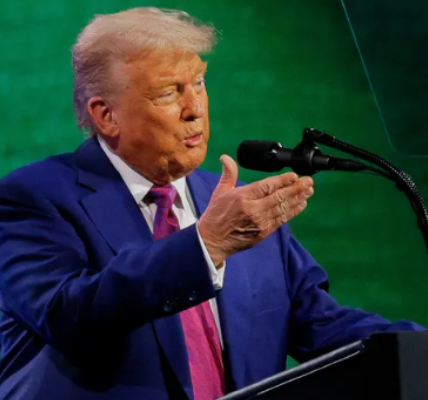 Donald Trump in a blue suit speaking into a microphone, gesturing with his right hand on stage with a green background.