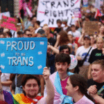 Crowd of over 100,000 people marching through central London streets during Trans+ Pride 2025, holding flags and signs in support of trans rights.