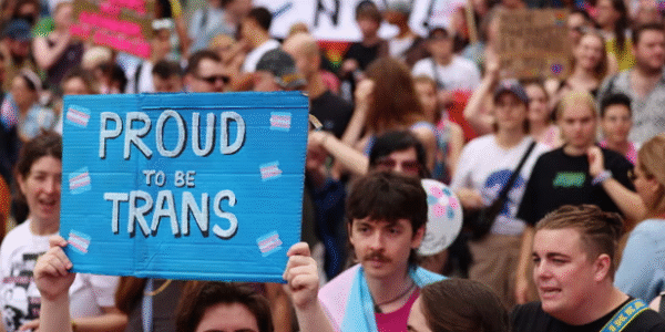 Crowd of over 100,000 people marching through central London streets during Trans+ Pride 2025, holding flags and signs in support of trans rights.